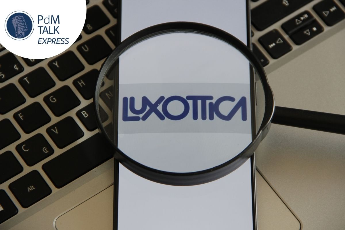 Logo Luxottica osservato con una lente d'ingrandimento, immagine di copertina della puntata di PdM Talk Express sul passaggio generazionale in EssilorLuxottica