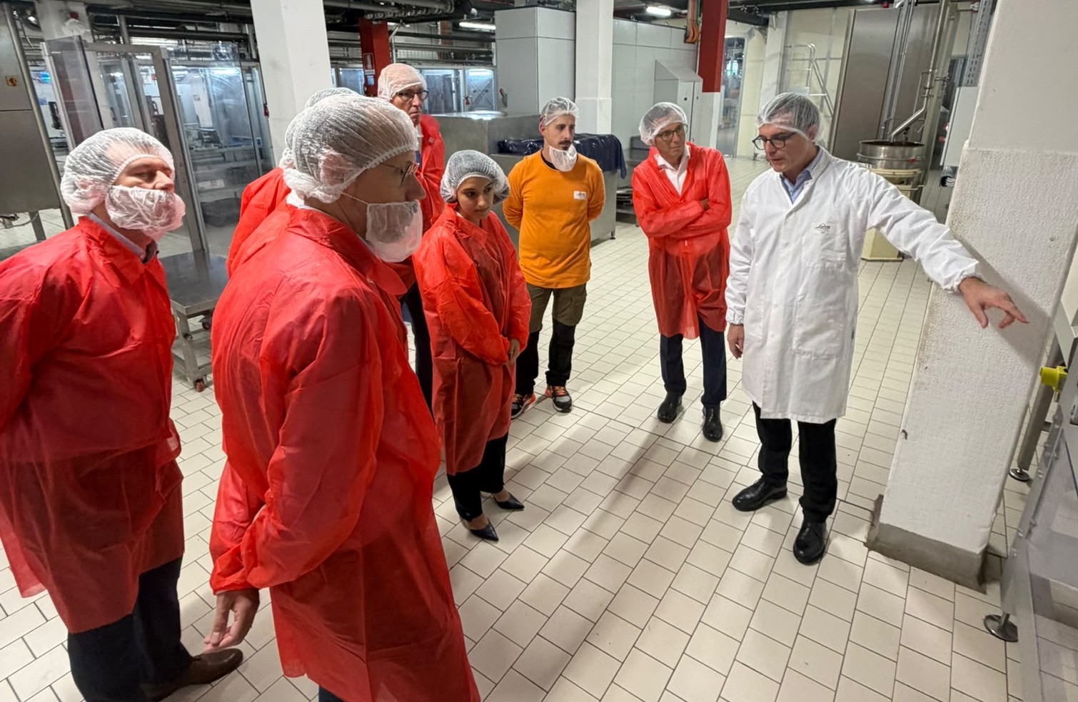Visita allo stabilimento Laica: un gruppo di partecipanti in camice rosso ascolta la spiegazione di un responsabile in camice bianco